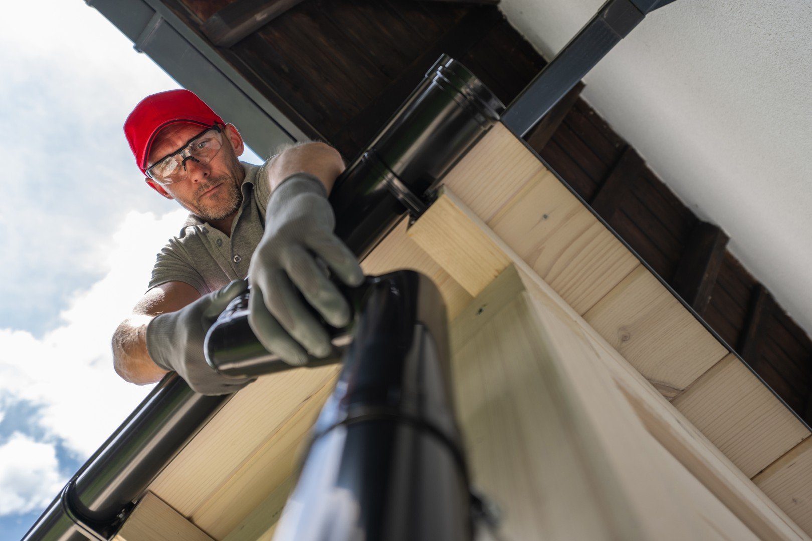 Man in a red cap and work gloves fitting a black downpipe below the eaves